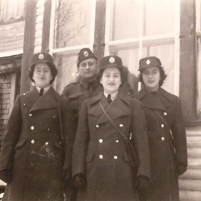 Photographie en noir et blanc – Trois jeunes femmes debout face à leur père, devant une maison. Elles portent toutes un uniforme militaire hivernal, et l'on aperçoit de la neige au sol.