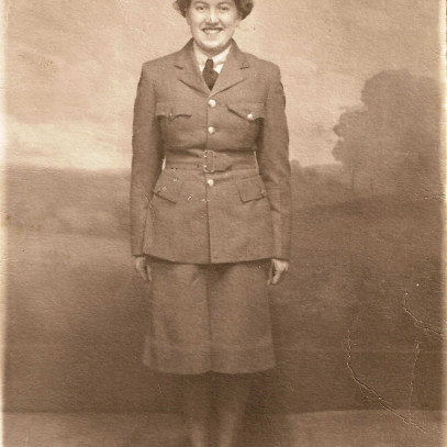 Photographie en tons sépia – Une jeune femme vêtue de son uniforme militaire complet, qui comporte une jupe. Elle est souriante, au garde-à-vous.