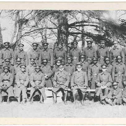 Photographie en noir et blanc – De nombreux hommes noirs sont assis pour une photo de groupe. La plupart portent de longs manteaux militaires. Des arbres et des toitures sont visibles à l’arrière-plan.