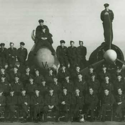 Photographie en noir et blanc – Une soixantaine de membres d’équipage du 404e escadron posent pour une photo de groupe devant un Beaufighter. Les hommes sont alignés en face de l’avion ou assis sur les moteurs ou les ailes.
