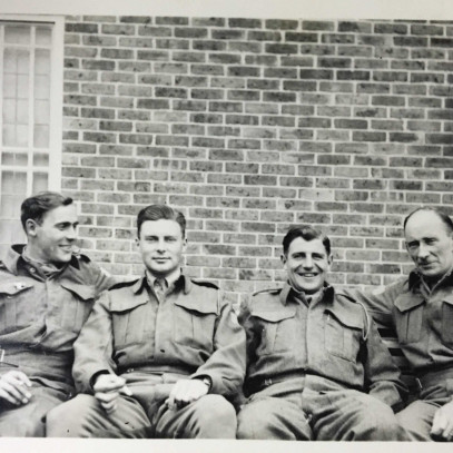 Photographie en noir et blanc – Archie, complètement à droite, est assis sur un banc avec trois hommes beaucoup plus jeunes. Tous sont en uniforme. Ils sourient.