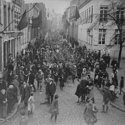 Photographie en noir et blanc – Des soldats canadiens jouant de la cornemuse défilent sur une rue envahie par la foule, approchant d'un espace découvert. Des drapeaux français sont accrochés aux édifices bordant la rue.