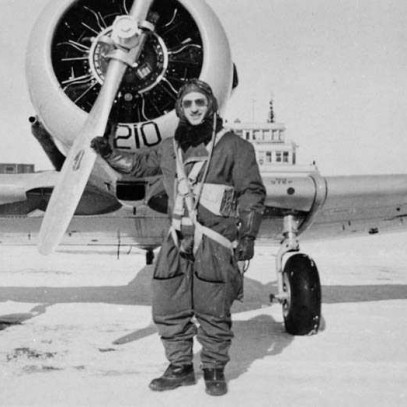 Photographie en noir et blanc – Un homme portant un lourd équipement de pilote se tient devant un avion, un bras déposé sur l’hélice.