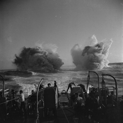 Photographie en noir et blanc – La poupe d’un immense navire; des hommes font dos à l’objectif de l’appareil photo. Au loin, des colonnes d’eau soufflent violemment à mesure qu’explosent des grenades sous-marines.