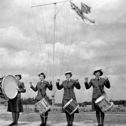 Photographie en noir et blanc – Quatre femmes en uniforme de la marine sont placées en rang. Celle à gauche joue de la grosse caisse. Les autres, à leur caisse claire, tiennent leurs baguettes à l’horizontale. On aperçoit derrière le Red Ensign.