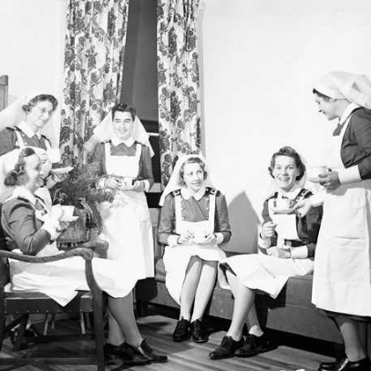 Photographie en noir et blanc – Six infirmières militaires (uniforme bleu avec un tablier et une coiffe de couleur blanche) sont assises ou debout. On voit une partie du mobilier en bois et des rideaux au motif floral. Elles bavardent en buvant du thé.