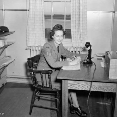 Photographie en noir et blanc – Une femme en tenue militaire est assise à un pupitre en bois dont la surface est couverte de feuilles. On aperçoit une petite étagère et un microphone ainsi que des tablettes et, à gauche, une fenêtre.