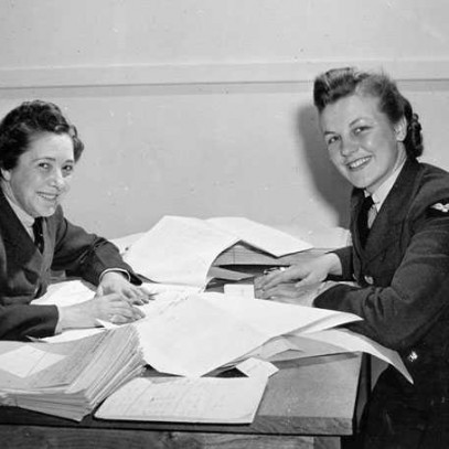 Photographie en noir et blanc – Deux femmes portant un uniforme de l’Aaviation royale du Canada, mais sans casquette, sont assises l’une en face de l’autre à une table couverte de piles de feuilles.