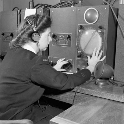 Photographie en noir et blanc – Une femme en tenue militaire, bien coiffée et aux ongles manucurés, regarde un relevé sur de l'équipement, utilisant une main pour régler un cadran. Assise à un bureau en bois, elle est entourée de machines similaires.