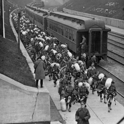 Photographie en noir et blanc – Des centaines de militaires portant un kilt marchent près du train dont ils ont débarqué. La voie ferrée se trouve entre des collines de verdure délimitées par un mur en ciment; un journaliste prend des notes ou une photo.