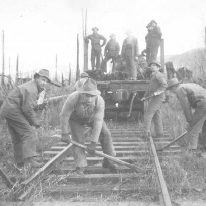 Photographie en noir et blanc – Cinq hommes en train de construire ou de réparer un chemin de fer à l’aide d’outils à long manche. Cinq autres les regardent à l’arrière d’une camionnette. Ils se trouvent dans une zone forestière.