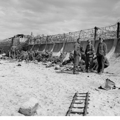 Photographie en noir et blanc – Une plage sablonneuse et une digue élevée, courbée et surmontée d'épais fils barbelés. Un nid de mitrailleuses est visible un peu plus loin sur la plage. Des prisonniers allemands sont assis contre la digue.