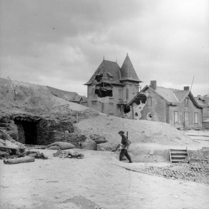 Photographie en noir et blanc – Des bâtiments en pierre sont visibles à l’arrière-plan, de même que de gros trous d’obus. Un soldat canadien solitaire marche vers un bunker couvert de sacs de sable, bâti sous une grosse dune.