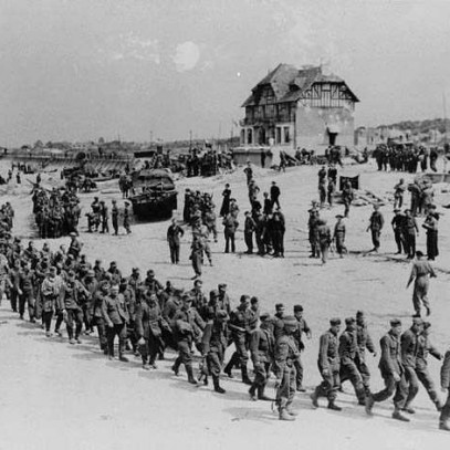 Photographie en noir et blanc – Une longue colonne de soldats allemands avançant sur une plage sablonneuse, sous le regard de militaires canadiens. Des obstacles de plage et des véhicules militaires sont visibles. La Maison du Canada surplombe la scène.