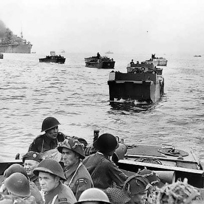 Photographie en noir et blanc – Une série de péniches de débarquement s’éloigne d’un immense navire. Des soldats contemplent la haute mer.