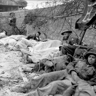 Photographie en noir et blanc – Des soldats blessés sont étendus près d’un muret de pierre sur la plage. Certains sont complètement ou partiellement couverts de draps blancs, alors que d’autres sont appuyés sur leurs coudes, fixant la caméra.