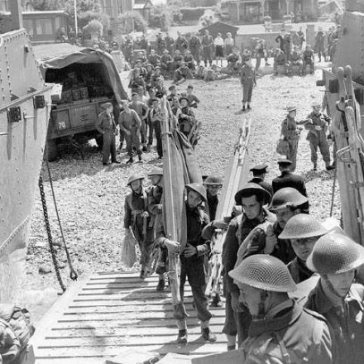 Photographie en noir et blanc – Des hommes en tenue de combat transportant de l’équipement (civières, etc.) avancent vers la rampe d’une péniche de débarquement. On aperçoit derrière d’autres hommes les observant. Il y a un camion militaire.
