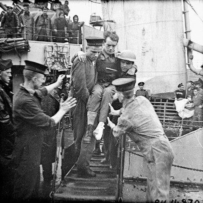 Photographie en noir et blanc – Un marin et un soldat transportent un soldat blessé vers la planche d’embarquement d’un grand bateau où deux autres marins attendent de le prendre à leur tour. D’autres hommes observent la scène du haut de ponts supérieurs.