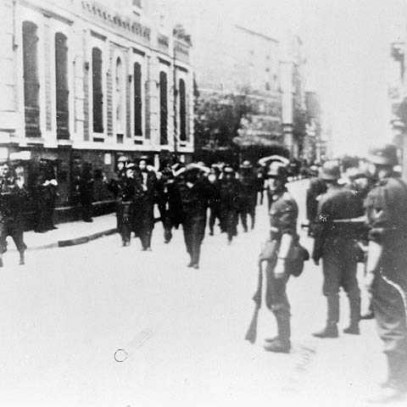 Photographie en noir et blanc – Des soldats canadiens marchent à travers les rues de Dieppe, les mains derrière la tête. Des soldats allemands armés, sur le côté, les surveillent.