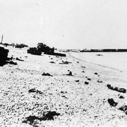Photographie en noir et blanc – Un quai est visible au loin. Des chars d’assaut sont abandonnés et des corps reposent sur la plage rocheuse.