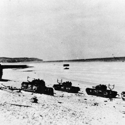 Photographie en noir et blanc – Quatre chars d’assaut et une péniche de débarquement sont abandonnés sur les plages rocheuses de Dieppe. Au loin, on aperçoit de l’équipement militaire reposant dans la mer à marée basse.