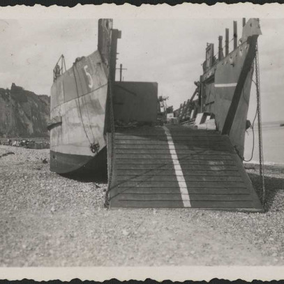 Photographie en noir et blanc – Une barge de débarquement de chars d’assaut sur une plage de galets à Dieppe. On aperçoit au loin les falaises blanches emblématiques de la région. La péniche se trouve entièrement sur la plage, et sa plateforme d’embarquem