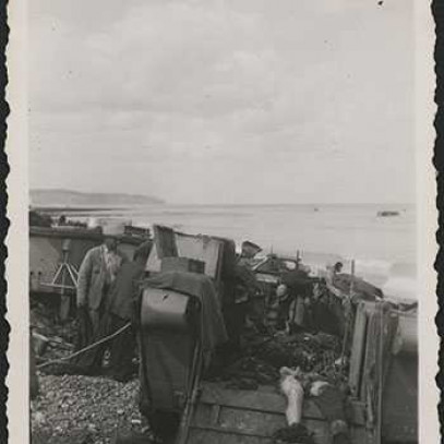 Photographie en noir et blanc – Des civils de Dieppe sont montrés en train de charger les cadavres de soldats canadiens sur un véhicule sur la plage. On aperçoit l’eau à l’arrière-plan.