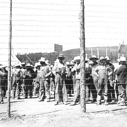 Photographie en noir et blanc – De nombreux hommes vêtus d’une salopette et d’un chapeau à large bord se trouvent derrière une haute clôture de barbelés. Ils ne font rien. Une tente est visible à l’arrière-plan.