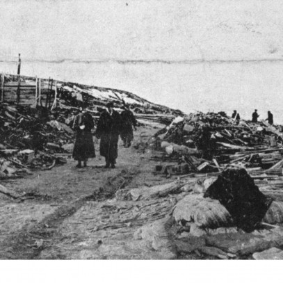Photographie en noir et blanc – Des survivants avancent le long d’un étroit sentier à travers les débris, loin du rivage. Le squelette d’un immense édifice est visible à gauche.