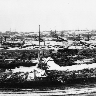 Photographie en noir et blanc – Une voie ferrée au premier plan; quelques structures visibles au loin. On aperçoit la fondation et les décombres d’un bâtiment au centre de la photo, de même que des arbres calcinés et de la neige au sol.