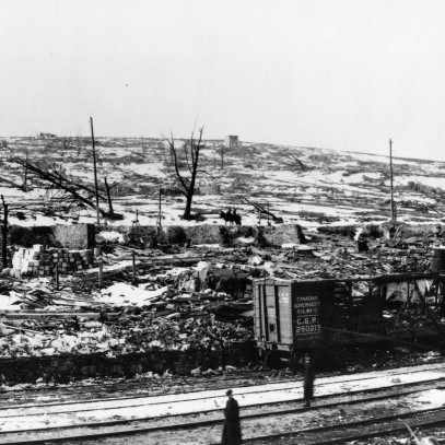 Photographie en noir et blanc – La voie ferrée au premier plan. La neige, les décombres, des arbres calcinés et des ruines de bâtiments composent le paysage. Un détail notable : un véhicule ferroviaire complètement détruit.