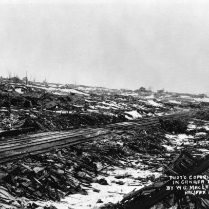 Photographie en noir et blanc – Les voies ferrées se prolongent au loin. On aperçoit un bâtiment soufflé; encore debout, il semble squelettique. Les décombres et les débris sont omniprésents. Les arbres sont calcinés et brisés.