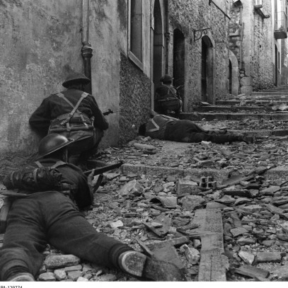 Photographie en noir et blanc – Des soldats canadiens en position accroupie ou couchée pointent leur fusil. Un homme gît inerte sur le sol près d'eux.
