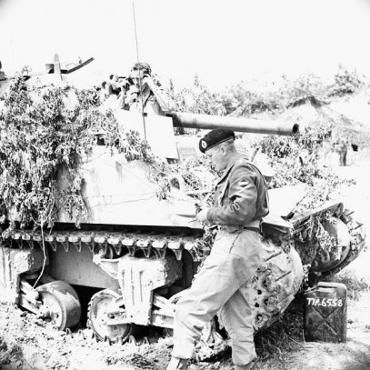 Photographie en noir et blanc – Un char d’assaut est partiellement couvert de branchages pour le camoufler. Un soldat jette un coup d’œil sur le dessus, alors qu’un autre reste devant. Un bidon d’essence est visible au sol.