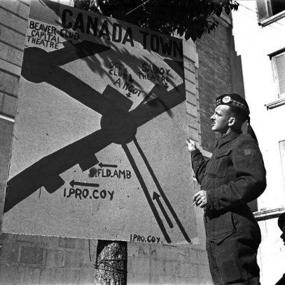 Photographie en noir et blanc – Un soldat pose devant un panneau sur lequel on peut lire « Canada Town » (« Canadaville). Celui-ci donne la direction vers divers lieux de divertissement dans la région.