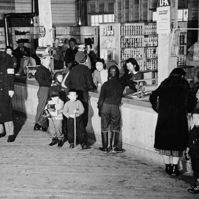 Photographie en noir et blanc – Des gens se trouvent devant et derrière le comptoir d’un magasin. Les tablettes sont remplies de boîtes de conserve et d’autres produits. Les gens conversent de part et d’autre du comptoir.