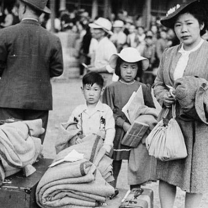 Photographie en noir et blanc – Une femme est debout avec ses deux enfants, endimanchés. On aperçoit tout près un amoncellement de biens personnels et de valises.