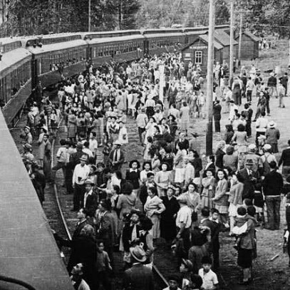 Photographie en noir et blanc – Un train de voyageurs à une gare. Une foule nombreuse est rassemblée. Des personnes parlent aux passagers à bord.