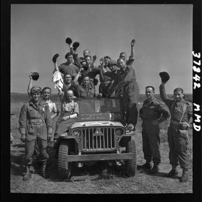 Photographie en noir et blanc – Un groupe d’hommes à l’intérieur et autour d’un tout-terrain militaire au milieu d’un champ vide; la plupart lèvent leur casquette pour saluer la caméra.