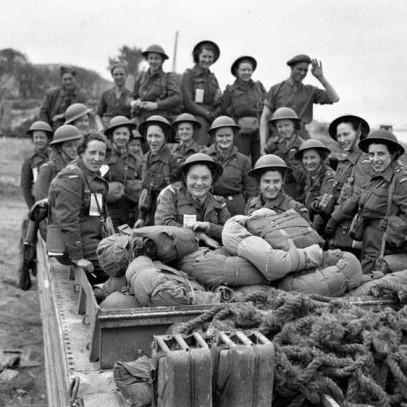 Photographie en noir et blanc – Des infirmières à l’arrière d’un véhicule militaire près d’une tête de pont sur une plage en Normandie. Elles sont coiffées de casques de métal.