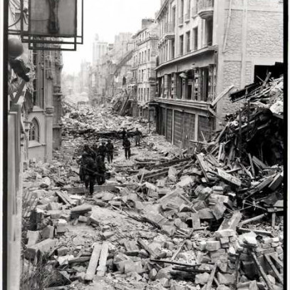 Photographie en noir et blanc – Des troupes canadiennes en rangée descendent le long d’une route étroite à Caen. Les bâtiments de part et d’autre montrent de nombreux dommages. Les rues sont complètement couvertes de gros morceaux de gravats.