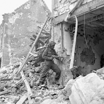 Photographie en noir et blanc – Un soldat se trouve sur un immense tas de décombres. Il pointe une arme vers un trou béant sur la devanture d’un bâtiment. Un autre immeuble en ruines est visible derrière lui, et la rue est entièrement couverte de débris.