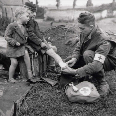 Photographie en noir et blanc – Un soldat (avec un brassard médical) bande la cheville d’un garçon sous le regard attentif d’un autre enfant.