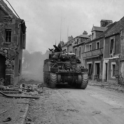 Photographie en noir et blanc – Les bâtiments d’une rue sont lourdement endommagés. Un char d’assaut Sherman se fraie un chemin à travers les décombres.