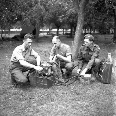 Photographie en noir et blanc – Trois soldats sont accroupis au sol, en train de travailler à une petite pièce de machinerie.
