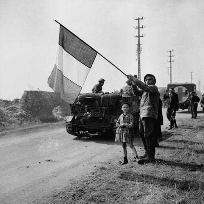 Photographie en noir et blanc – Un Français avec un garçonnet en bordure de route tient un grand drapeau de la France. Des soldats défilent à pied et dans des véhicules militaires.