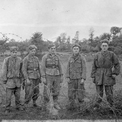 Photographie en noir et blanc – Cinq très jeunes soldats allemands en diverses tenues militaires se trouvent derrière une épaisse clôture de fil barbelé.
