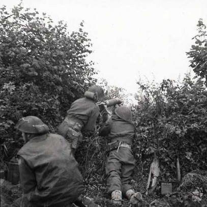 Photographie en noir et blanc – Trois soldats sont accroupis dans des buissons épais. Deux dirigent une arme vers un passage étroit. Le troisième regarde de côté. Sur le sol se trouvent de l’équipement, des armes et des munitions.