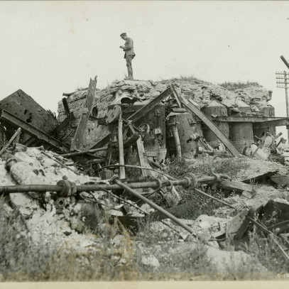 Photographie en noir et blanc – Un soldat se trouve sur les vestiges d’une raffinerie de sucre. Diverses pièces mécaniques sont visibles dans les débris.
