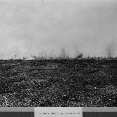 Photographie en noir et blanc – La lueur des explosions d’obus est visible au loin au-dessus du no man’s land stérile (au bas de l’image). Le sol est boueux et inégal; des arbres morts sont dispersés ça et là..